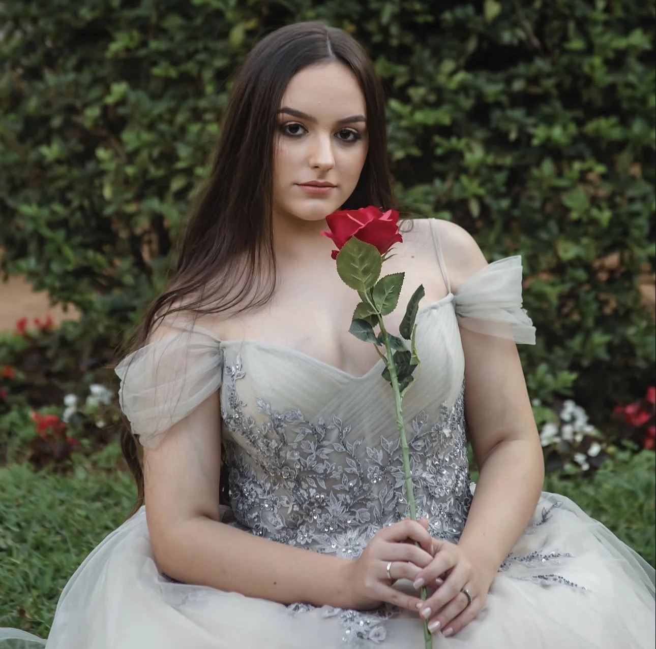 Jovem com vestido claro segura uma rosa vermelha diante de um jardim