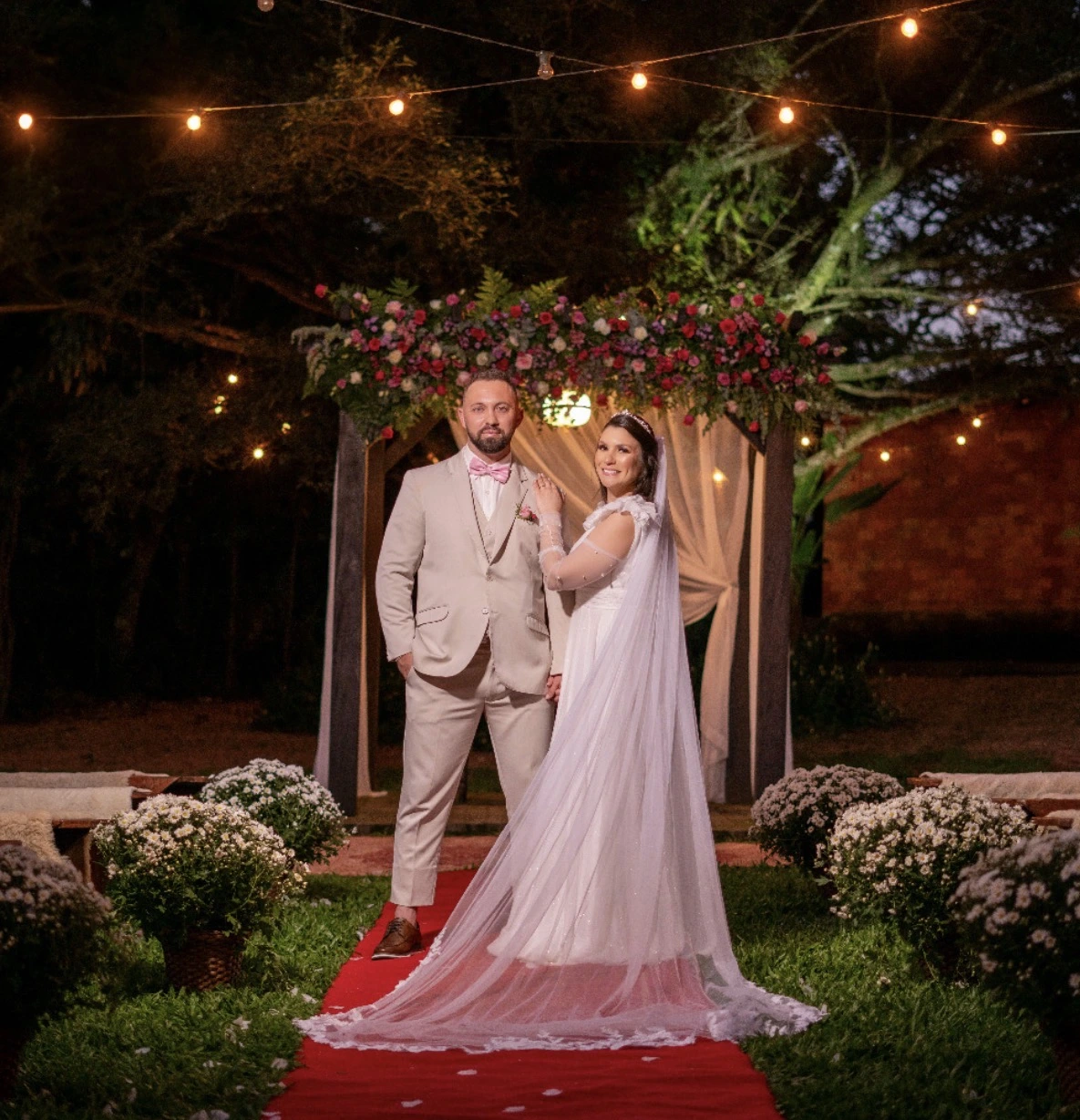 Casal de noivos sob luzes de jardim à noite, com arco de flores